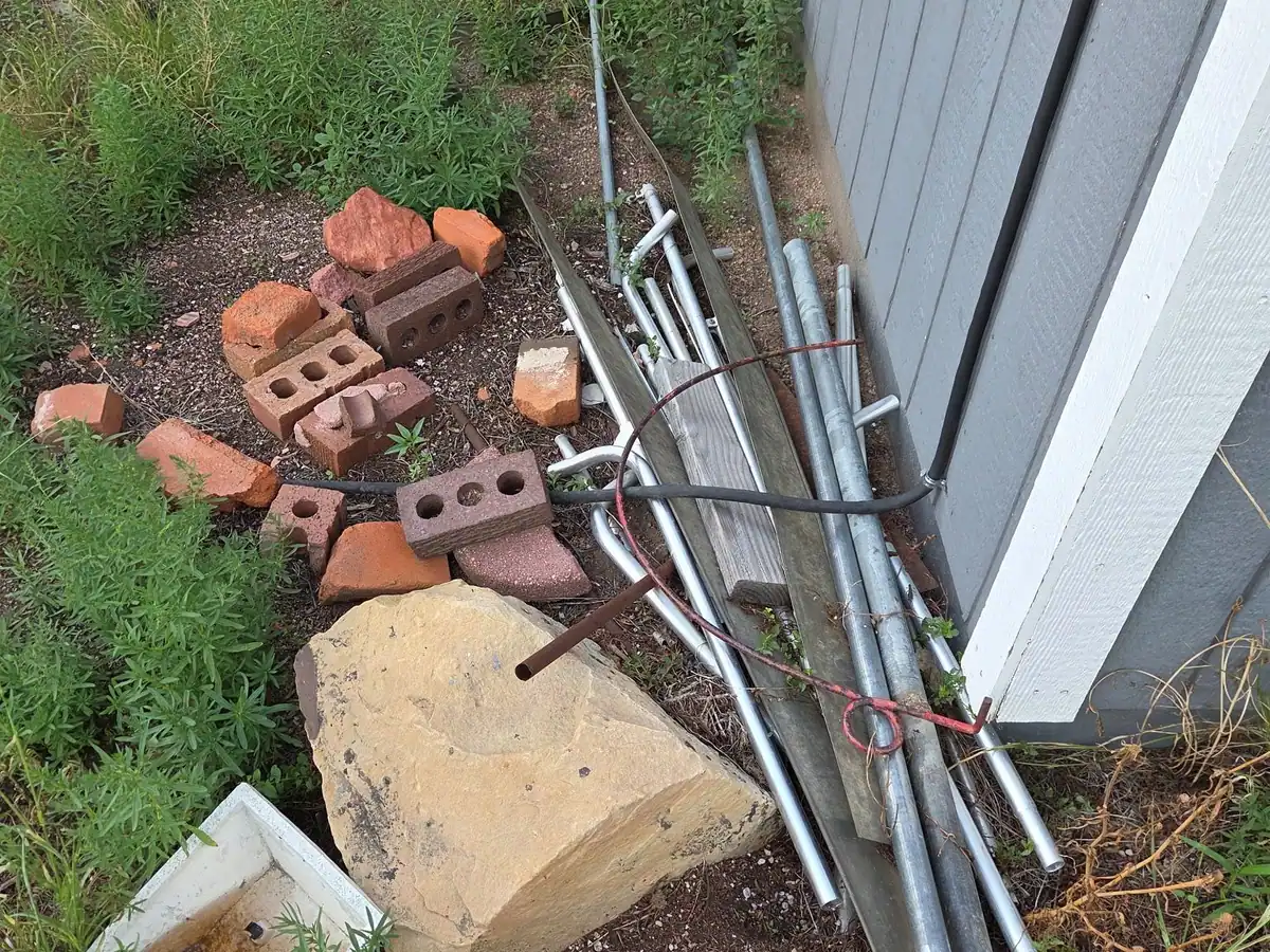 Broken bricks and old metal poles piled in the weeds beside a grey shed