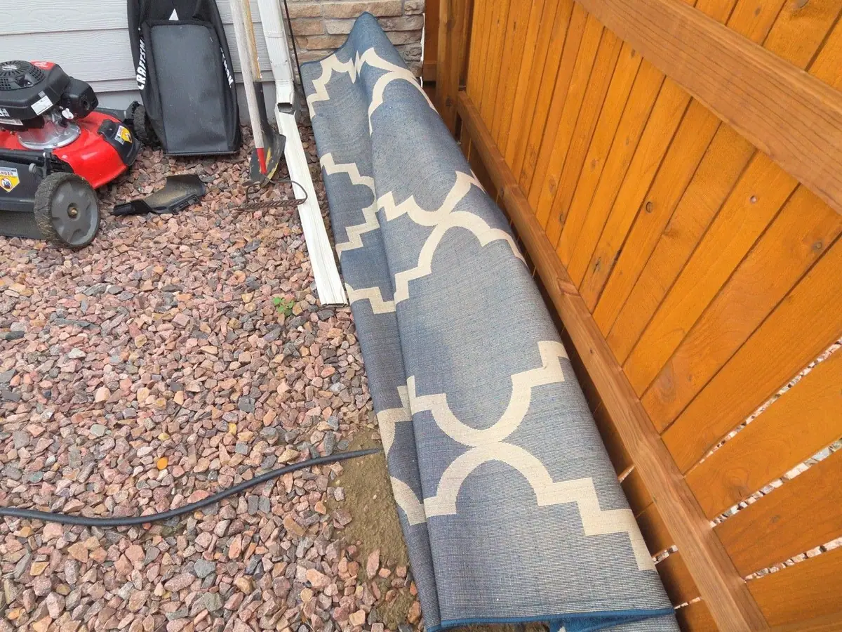 A rolled rug and an old mower against a wood fence in a side yard